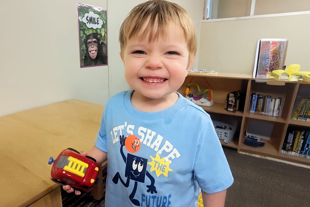Child indoors smiling widely while holding a red toy car.